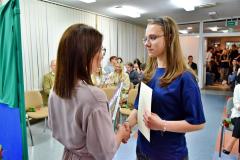 Zbliżenie na moment wręczania nagrody. Wychowawczyni (widoczna z profilu) podaje rękę uśmiechniętej wychowance. Dziewczyna trzyma w drugiej ręce dyplom. W tle widać siedzących na krzesłach pozostałych uczestników uroczystości.