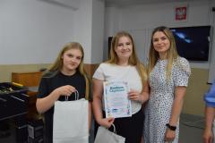 Dwie uśmiechnięte wychowanki stoją wraz wychowawczynią. Dziewczyna pośrodku dumnie pokazuje dyplom z kolorowym napisem „Konkurs Czystości”. Obie wychowanki trzymają w rękach białe, papierowe torby z upominkami.