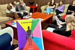 Wychowawca trzyma w dłoni kolorową ulotkę kampanii „W pełnym wymiarze” Polskiej Akcji Humanitarnej. W tle grupa młodzieży siedząca na kanapach i fotelach, zaangażowana w przeglądanie materiałów edukacyjnych. Scena przedstawia atmosferę dyskusji i refleksji nad tematami społecznymi poruszanymi w ramach akcji.