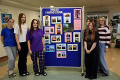 Sześcioro wychowanek bursy stoi przy tablicy przedstawiającej wystawę fotografii. Na zdjęciach i obrazie znajdują się elementy przyrody: drzewa, liście, niebo oraz elementy krajobrazu miejskiego: budynki, most, droga. 