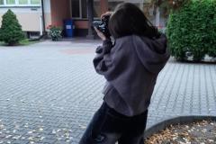 Wychowanka stoi przed budynkiem bursy i wykonuje zdjęcie aparatem fotograficznym.