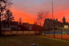 Na pierwszym planie widnieje boisko bursy. W tle zachód słońca, drzewa, budynki.