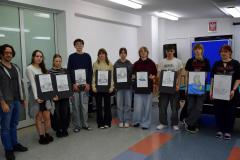 Dziewięcioro wychowanków bursy stoi w pół okręgu. Obok wychowanków stoi artysta malarz. W dłoniach trzymają kartki z namalowaną przez nich (na warsztatach malarskich) martwą naturę: dzbanek stojący na materiale, obok leży cytryna.