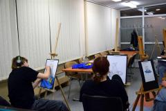 Dwie wychowanki siedzą przy sztalugach malarskich. Na kartkach rysują ołówkiem i pastelami martwą naturę (dzbanek który stoi na materiale, przy nim leży cytryna). W oddali widać krzesła i stoły na których stoją dwie ekspozycje: dzbanek na niebieskim i granatowym materiale, z boku leży przekrojona cytryna.