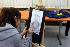 Wychowanka siedzi przy sztaludze malarskiej. Na kartce rysuje ołówkiem martwą naturę (dzbanek który stoi na granatowym materiale, przy nim leży przekrojona na pół cytryna).