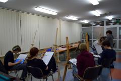 Dziewięcioro wychowanków siedzi przy sztalugach malarskich. Na kartkach rysują ołówkiem i pastelami martwą naturę (dzbanek który stoi na materiale, przy nim leży przekrojona na pół cytryna). W oddali widać krzesła i stoły na których stoją dwie ekspozycje: dzbanek na niebieskim i granatowym materiale, z boku leży przekrojona cytryna. Przy wychowankach stoi Pan prowadzący warsztaty pokazujący i tłumaczący wychowankom sposoby malowania.