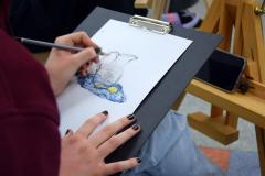 Wychowanka siedzi przy sztaludze malarskiej. Na kartce rysuje ołówkiem martwą naturę (dzbanek który stoi na granatowym materiale, przy nim leży przekrojona na pół cytryna).