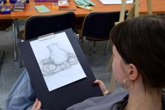 Wychowanka siedzi przy sztaludze malarskiej. Na kartce rysuje ołówkiem martwą naturę (dzbanek który stoi na granatowym materiale, przy nim leży przekrojona na pół cytryna). W oddali na stołach leżą artykuły plastyczne: pastele, kredki ołówki.