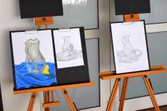 W holu bursy stoją dwie sztalugi malarskie. Na sztalugach stoi pięć prac. Do czarnych podkładek przypięte są białe kartki na których znajduje się namalowana ołówkiem lub pastelami martwa natura (dzbanek który stoi na materiale, przy nim leży przekrojona na pół cytryna)