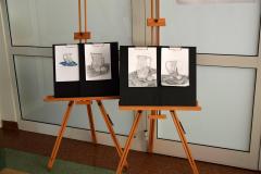 W holu bursy stoją dwie sztalugi malarskie. Na sztalugach stoją cztery prace. Do czarnych podkładek przypięte są białe kartki na których znajduje się namalowana ołówkiem lub pastelami martwa natura (dzbanek który stoi na materiale, przy nim leży przekrojona na pół cytryna)