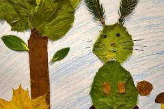 Na zdjęciu widać zajączka wyklejonego z liści i igieł oraz drzewo wykonane z jesiennych liści na tle narysowanego krajobrazu