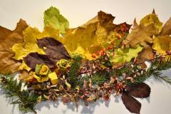 Na zdjęciu widać kompozycję z jesiennych liści, gałązek iglastych i czerwonych owoców berberysu ułożoną na białym tle