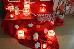 Na rozłożonym czerwonym płótnie stoją biało-czerwone lampiony i kotyliony patriotyczne. W lampionach palą się świece. 