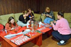 Pięć wychowanek siedzi przy stole, wykonując biało-czerwone kotyliony patriotyczne i lampiony. Na stole leżą artykuły plastyczne: bibuła, nożyczki, wykonane kotyliony, lampiony. 