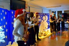 Na pierwszym planie stoi 10 osób: wychowankowie bursy. Ubrani odświętnie w biało – czarne stroje. Na głowie 5 osób znajdują się czapki Mikołaja. W rękach trzymają mikrofony i podkładki ze scenariuszami uroczystości. Wychowanka stojąca po lewej stronie trzyma w rękach saksofon. Za nimi znajduje się niebieska tablica na której znajdują się: wycięte z papieru białe gwiazdki, 2 białe anioły, lampki, 4 białe choinki. Pośrodku w drewnianym żłóbku leży postać Jezusa. Na tablicy znajduje się napis: Cicha noc, święta noc. Nad nim wisi biała duża gwiazda. Na podłodze znajduje się biała tkanina, na której leżą zapalone lampki.