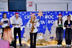 Na pierwszym planie stoi 5 osób: wychowankowie bursy. Ubrani odświętnie w biało – czarne stroje. Na głowie 3 osób znajdują się czapki Mikołaja. W rękach trzymają mikrofony i podkładki ze scenariuszami uroczystości. Wychowanka stojąca na środku trzyma w rękach saksofon, na którym gra kolędę. Za nimi znajduje się niebieska tablica na której znajdują się: wycięte z papieru białe gwiazdki, 2 białe anioły, lampki, 4 białe choinki. Pośrodku w drewnianym żłóbku leży postać Jezusa. Na tablicy znajduje się napis: Cicha noc, święta noc. Nad nim wisi biała duża gwiazda. Na podłodze znajduje się biała tkanina, na której leżą zapalone lampki.