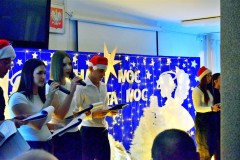 Na pierwszym planie stoi 5 osób: wychowankowie bursy. Ubrani odświętnie w biało – czarne stroje. Na głowie 2 osób znajdują się czapki Mikołaja. W rękach trzymają mikrofony i podkładki ze scenariuszami uroczystości. Dwie wychowanki stojące po lewej stronie trzymają w rękach mikrofony, śpiewają kolędę. Za nimi jest niebieska tablica na której znajdują się: wycięte z papieru białe gwiazdki, 2 białe anioły, lampki, 4 białe choinki. Pośrodku w drewnianym żłóbku leży postać Jezusa. Na tablicy znajduje się napis: Cicha noc, święta noc. Nad nim wisi biała duża gwiazda. Na podłodze znajduje się biała tkanina, na której leżą zapalone lampki.