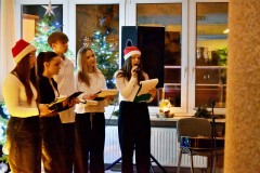 Pięcioro wychowanków stoi. W rękach trzymają podkładki i mikrofony. W tle stoją zapalone choinki. Wychowanka w czapce Mikołaj śpiewa kolędę. Na parapecie stoi zapalona świeca w szklanym wazonie.