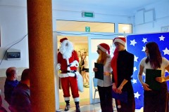 Do Sali wychowanek bursy w stroju świętego Mikołaja. W rękach trzyma dzwonek, za sobą worek z prezentami. Po prawej stronie stoi 3 wychowanków. W oddali (w holu bursy) widać stojące wychowanki bursy.