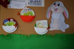 Na zdjęciu widać tablicę korkową ozdobioną wielkanocnymi dekoracjami: papierowymi jajkami z trawką oraz dużym, uśmiechniętym króliczkiem. Na dole znajduje się zielona bibuła imitująca trawę