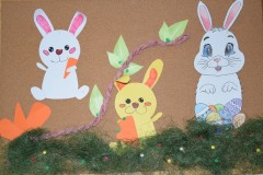 Na zdjęciu widać kolorową dekorację z papierowymi zajączkami. Trzy króliczki trzymają marchewki, siedzą na zielonej trawie, a obok nich są pisanki i gałązka z liśćmi