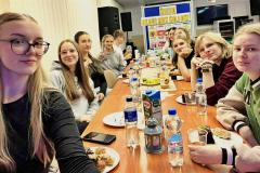 Młodzież siedząca przy długim stole w świetlicy bursy podczas spotkania z okazji Dnia Wolontariusza, wspólnie spędzająca czas przy przekąskach i napojach.