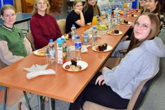 Grupa młodych osób siedzi przy stole w świetlicy bursy, jedząc przekąski i rozmawiając podczas spotkania z okazji Dnia Wolontariusza.