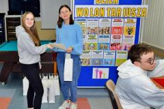 Podczas Dnia Wolontariusza wychowawczyni wręcza uczestniczce dyplom i upominek; w tle znajdują się plakaty informacyjne o wolontariacie.