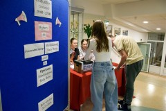 Komisja wyborcza wydaje karty do głosowania wychowankowi. Obok wychowanka wrzuca karty do głosowania do urny wyborczej. W tle komisja wyborcza oraz tablica z ogłoszeniem dotyczącej organizacji wyborów.