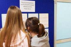 Dwie wychowanki obserwują umieszczone na tablicy ankiety kandydatów na przewodniczącego Młodzieżowej Rady Bursy.