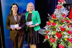 Dwie wychowawczynie bursy stoją na scenie z przypiętymi Medalami Komisji Edukacji Narodowej. W rękach trzymają legitymacje. W tle znajduje się baner oraz dekoracyjna aranżacja kwiatowa