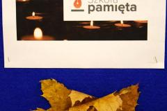 Napis: Pamiętamy – Odeszli do wieczności. W dłoniach znajduje się zapalony znicz, białe kwiaty i świeczki.