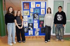 Czworo wychowanków bursy stoi przy tablicy przedstawiającej prace plastyczne: rysunki wykonane przez wychowanków. Rysunki przedstawiają: drzewa, kwiaty, ludzkie twarze, przyrodę.