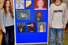 Dwoje wychowanków bursy stoi przy niebieskiej tablicy. Na tablicy znajduje się wystawa prac wychowanków (obrazów), na której widnieją elementy ludzkie (twarze, dłonie), zwierzęta, schody, zegar ścienny.