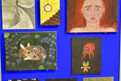 Na niebieskiej tablicy znajduje się wystawa prac wychowanków (obrazów), na której widnieją elementy ludzkie (twarze, dłonie), zwierzęta, schody, zegar ścienny.