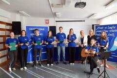 6 dziewcząt i 2 chłopców w niebieskich koszulkach z napisem Bursa Regionalna w Ostrołęce z  wychowawcami koordynującymi uroczystość zakończenia roku szkolnego pozują do zdjęcia. W tle widać tablice dekoracyjne z cytatem i żaglami.