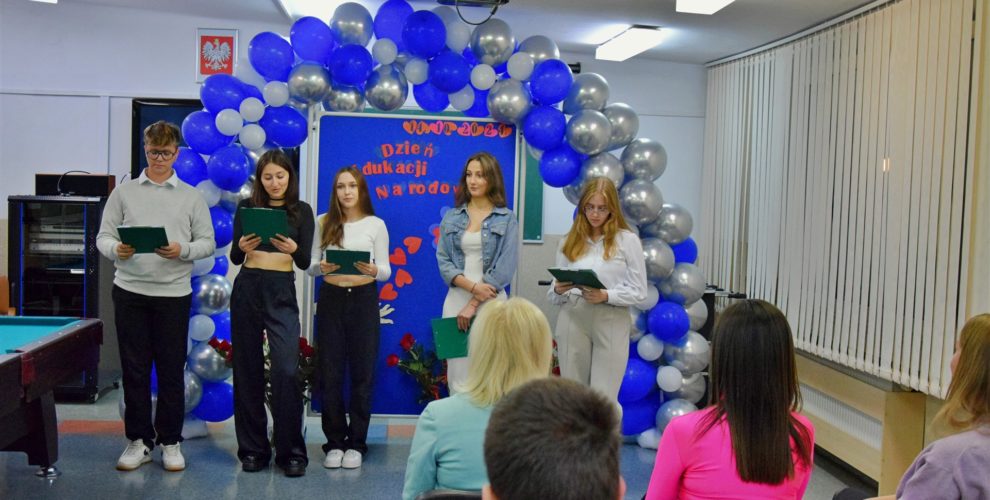Grupa wychowanków bursy stoi przed publicznością z tekstami w rękach, gotowa do występu artystycznego. W tle widoczna jest dekoracja z niebieskich i srebrnych balonów oraz tablica z napisem 'Dzień Edukacji Narodowej'. Na pierwszym planie siedzą widzowie.