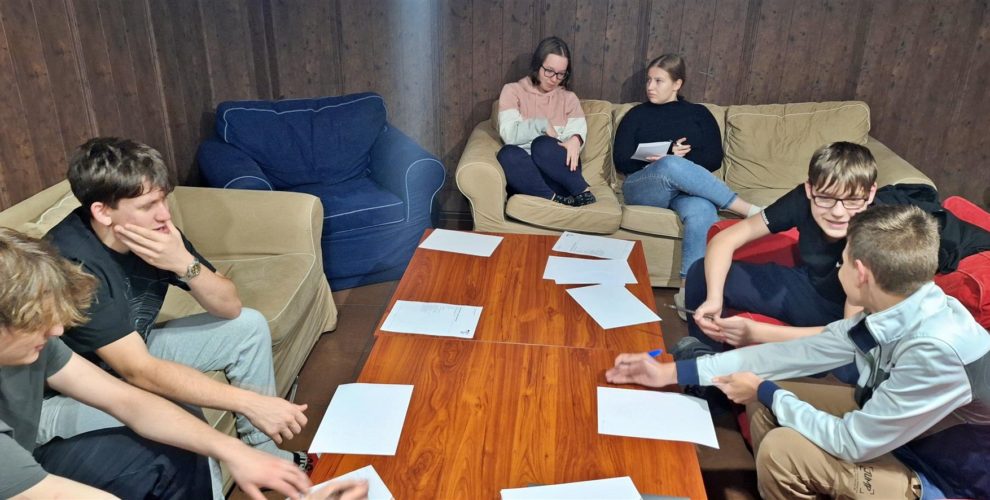 Kilka osób siedzi na kanapach i fotelach, pochylonych nad stołem zaangażowanych w rozmowę