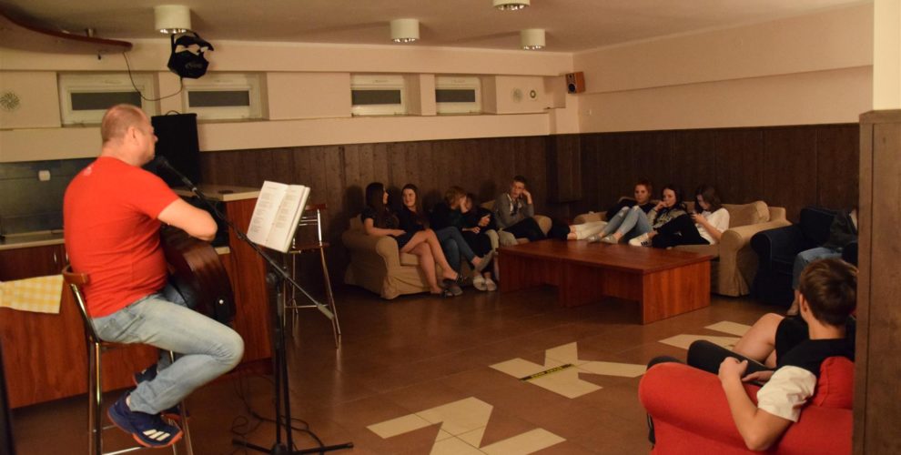 Wychowawca gra na gitarze, a wychowankowie siedzący na kanapach wokół, słuchają muzyki