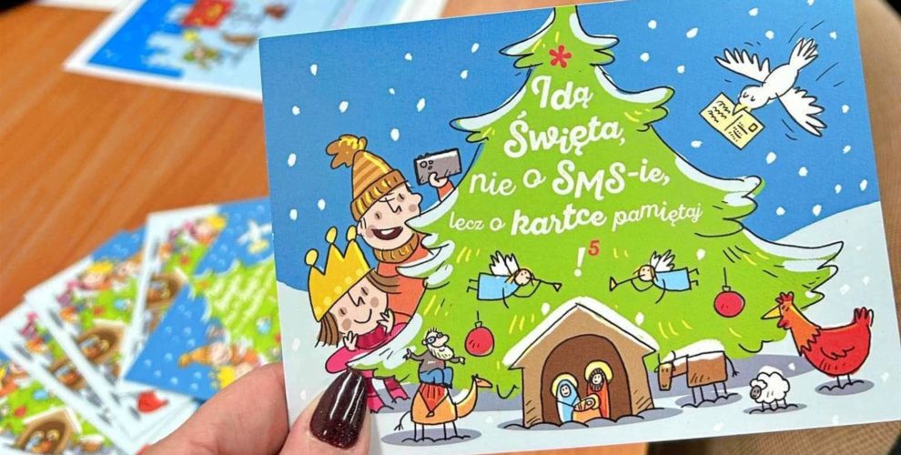 Na pierwszym planie znajduje się kartka z logo akcji. Młodzież siedzi przy stole i wypisuje karty świąteczne do różnych instytucji