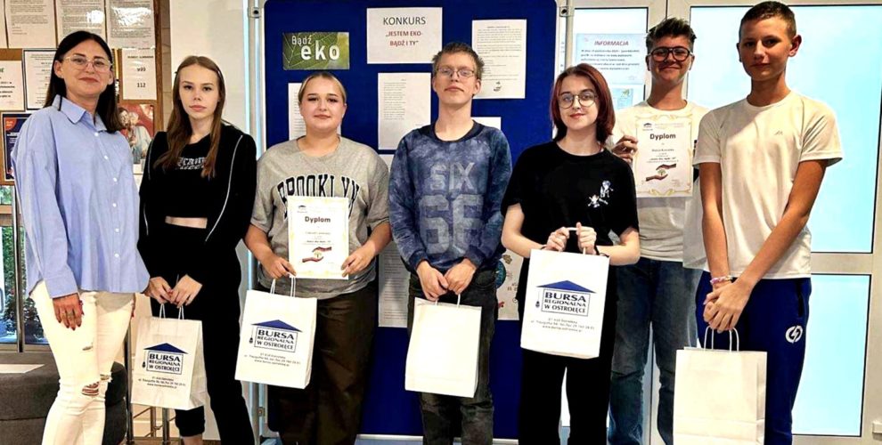 Wychowankowie biorący udział w konkursie stoją, trzymają w rękach nagrody oraz dyplomy. W tle znajduje się wystawa na której zaprezentowane są wiersze młodzieży.