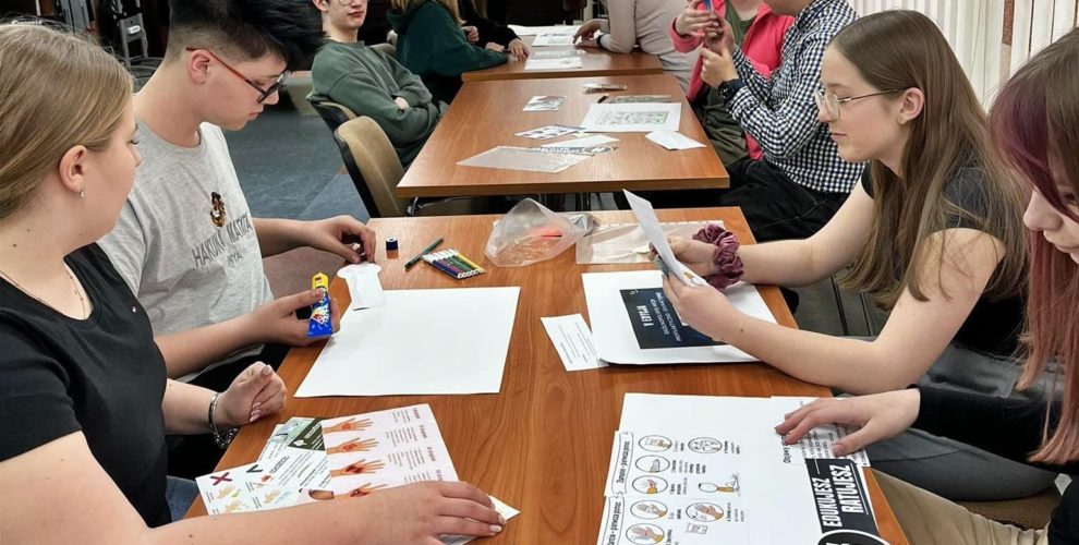 Grupa młodzieży uczestniczy w zajęciach edukacyjnych w ramach akcji „Edukujesz – Ratujesz”. Uczestnicy siedzą przy długim stole, pracując w parach lub małych grupach. Przed nimi znajdują się różne materiały dydaktyczne, takie jak ulotki, plakaty, arkusze papieru, flamastry, klej. Młodzież skupiona jest na przygotowywaniu materiałów związanych z pierwszą pomocą-niektórzy czytają instrukcje, inni wycinają elementy lub komponują plakaty.