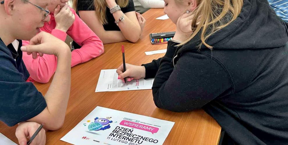 Grupa młodych osób siedzi przy stole i wspólnie pracuje nad zadaniem związanym z „Dniem Bezpiecznego Internetu”. Wychowankowie są skupieni, zapisują odpowiedzi na kartkach i korzystają z kolorowych materiałów edukacyjnych. Na pierwszym planie widoczna jest ulotka zatytułowana „Dekalog Cyfrowego Obywatela”, a w tle widać uczestników w swobodnej atmosferze pracy.