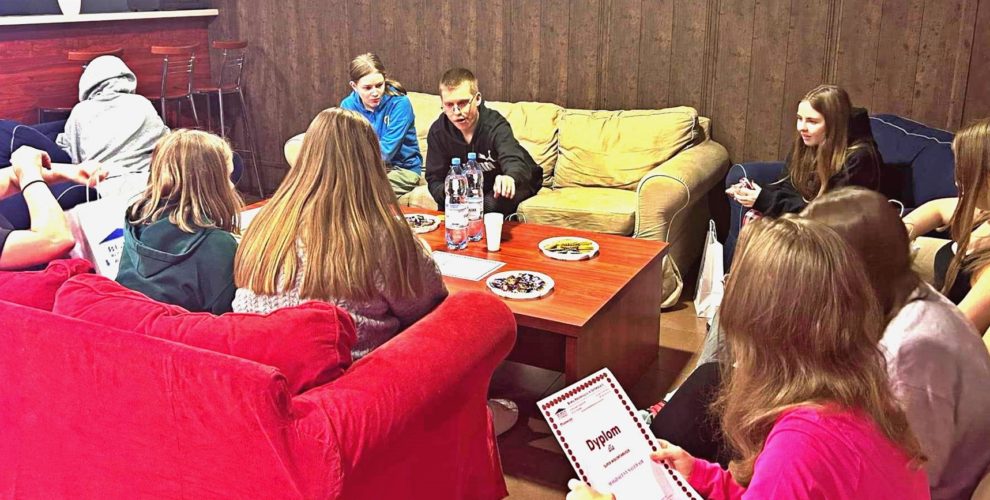 Młodzież siedzi na kanapach. Na stole znajduje się poczęstunek.