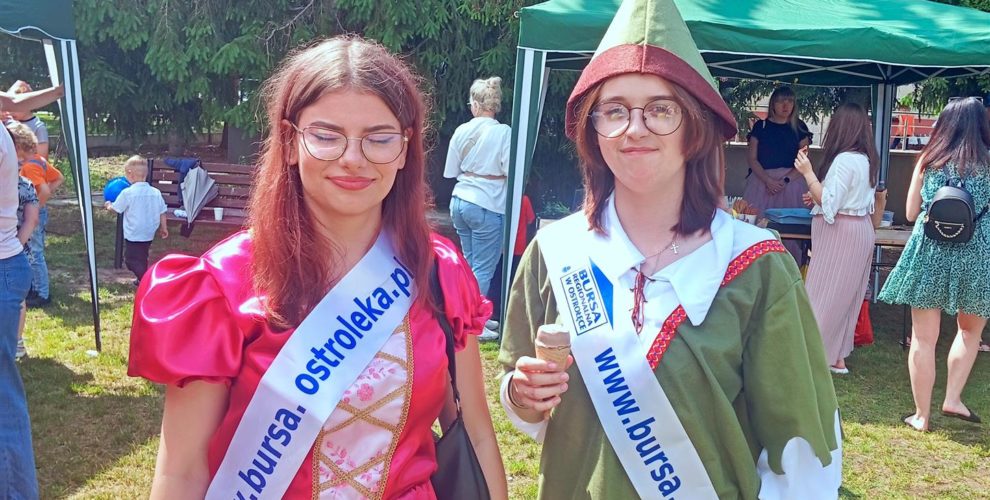 Wychowanki w strojach bajkowych postaci oraz wstęgą promującą bursę pozują na tle drzew oraz stanowisk ze słodkościami.