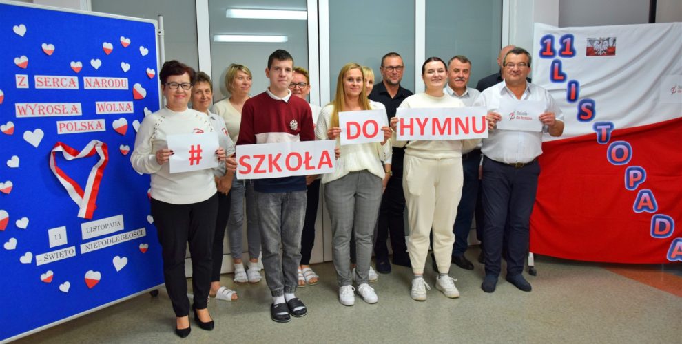 Grupa pracowników i uczniów bursy pozuje na tle patriotycznych dekoracji z okazji Święta Niepodległości. Trzymają tabliczki z hasłem „#SzkołaDoHymnu”.