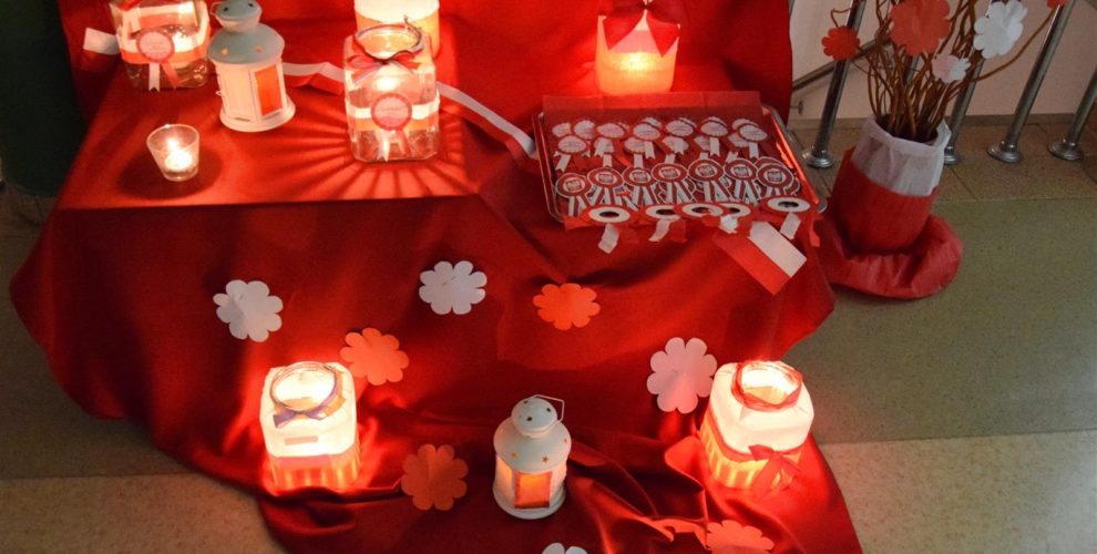 Na rozłożonym czerwonym płótnie stoją biało-czerwone lampiony i kotyliony patriotyczne. W lampionach palą się świece.