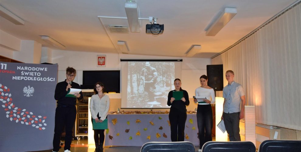 Młodzież w skupieniu prowadzi wieczór patriotyczny, w tle tablica z napisem 11 listopada, Narodowe Święto Niepodległości, Białym Orłem i liśćmi w kolorze biało czerwonym oraz dekoracja z liśćmi jesiennymi , wśród których stoją zapalone lampiony.