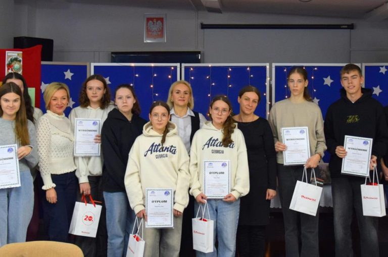 Na zdjęciu znajdują uczestnicy XIII Międzyinternackiego konkursu plastycznego na „Kartkę Bożonarodzeniową” wraz z opiekunami. W dłoniach trzymają dyplomy i nagrody. Z tyłu znajduje się czerwona tablica z kartkami konkursowymi i niebieska tablica z migającymi światełkami i białymi gwiazdkami.