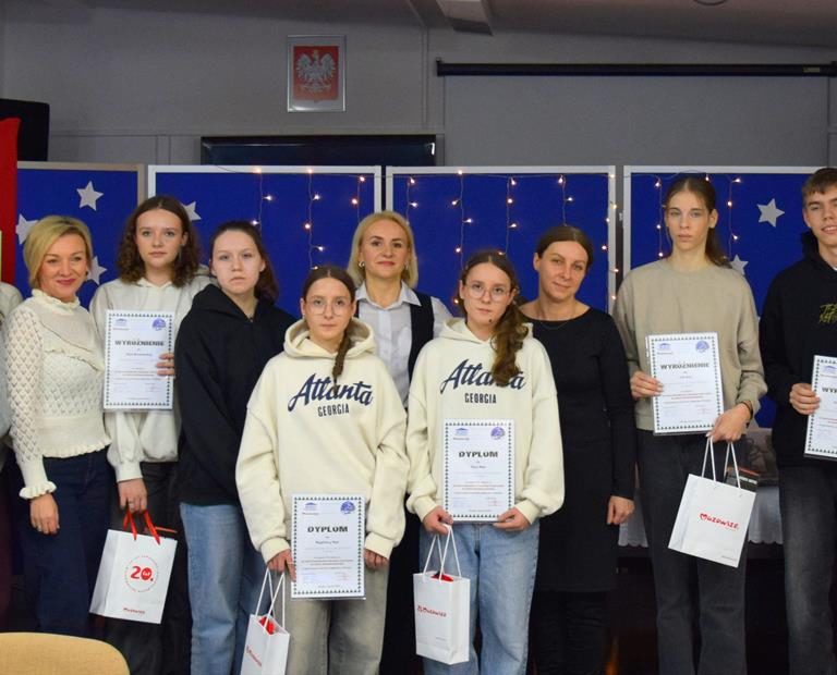 Na zdjęciu znajdują uczestnicy XIII Międzyinternackiego konkursu plastycznego na „Kartkę Bożonarodzeniową” wraz z opiekunami. W dłoniach trzymają dyplomy i nagrody. Z tyłu znajduje się czerwona tablica z kartkami konkursowymi i niebieska tablica z migającymi światełkami i białymi gwiazdkami.